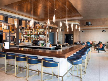 The image shows a modern bar with a central counter, blue-and-gold barstools, hanging lights, and a well-stocked back bar shelf.