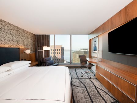 The image shows a modern hotel room with a large bed, TV, desk, and floor-to-ceiling window with a city view.
