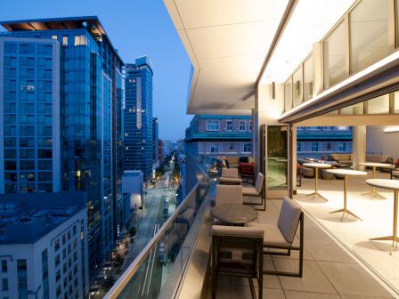 A modern balcony with seating overlooks a cityscape at dusk, with skyscrapers and illuminated streets visible.
