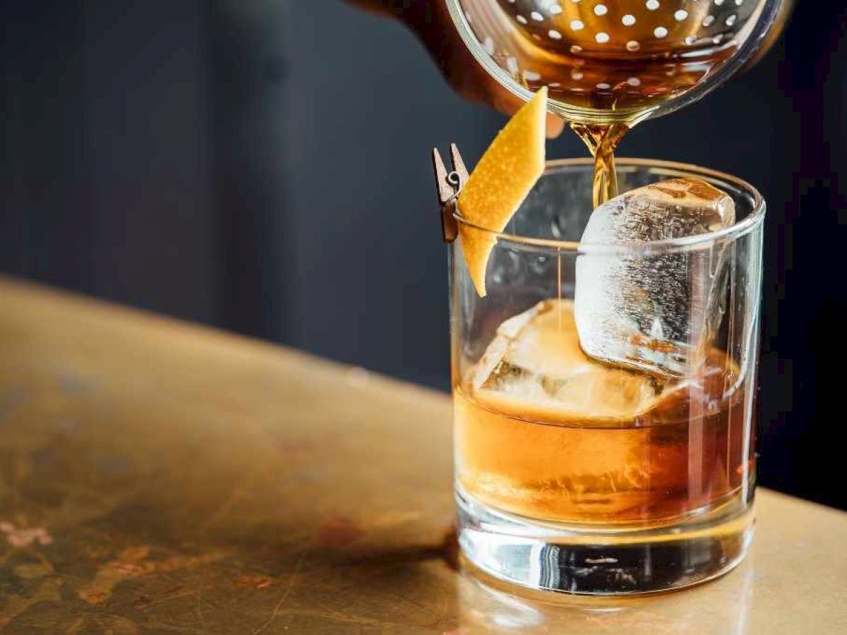 A bartender pours a drink over ice with an orange peel garnish in a glass on a bar table.