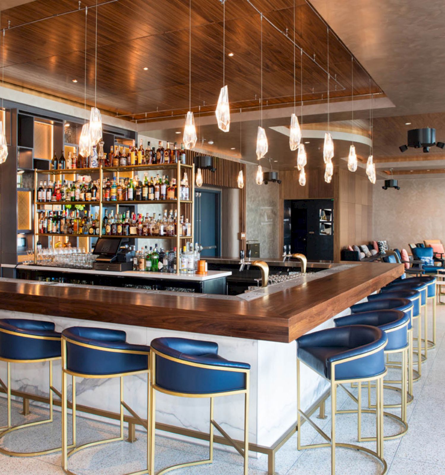 The image shows a modern bar with blue cushioned stools, a well-stocked bar, pendant lighting, and a spacious, stylish interior design.
