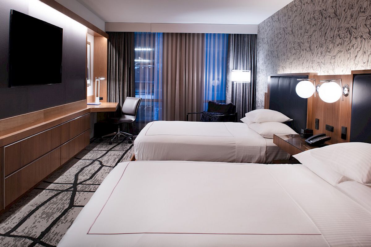 A modern hotel room with two beds, a large TV, desk area, and stylish lighting.