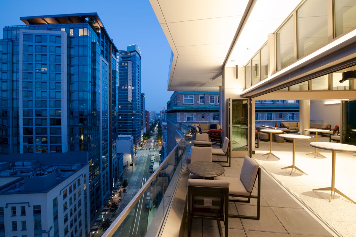 A modern urban balcony with seating overlooks a city street lined with tall buildings at dusk.