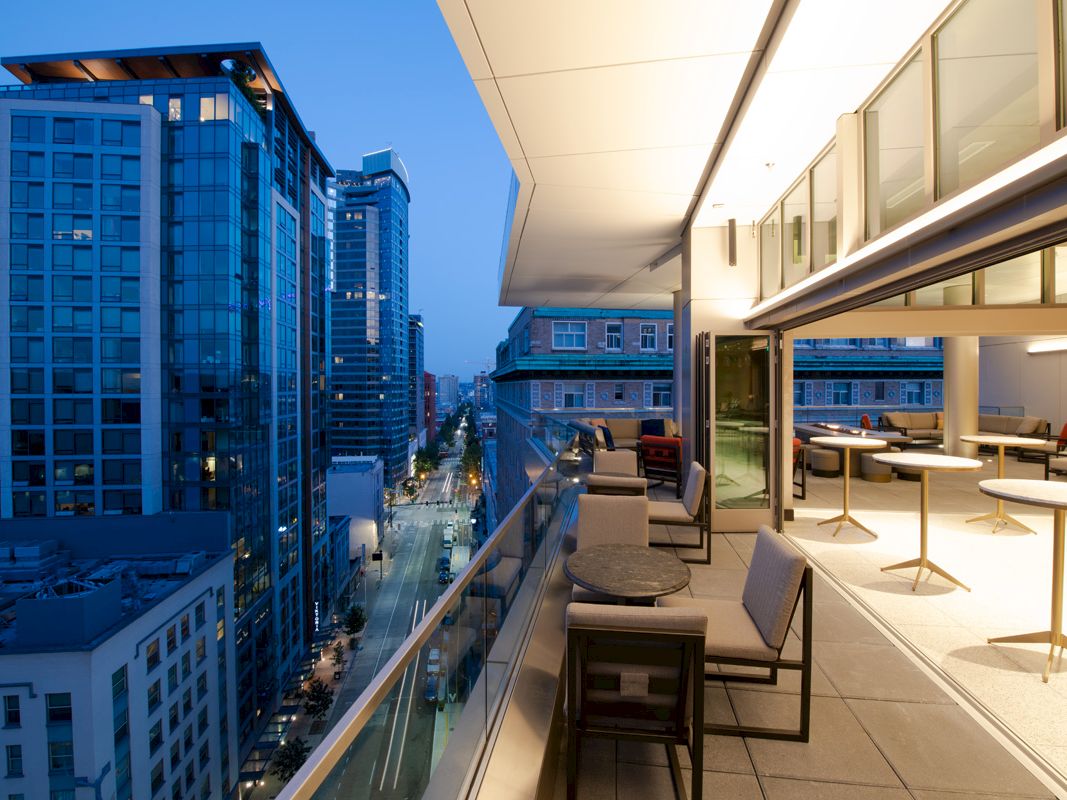A modern urban balcony with seating overlooks a city street lined with tall buildings at dusk.