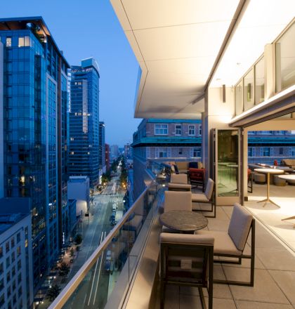 A modern city balcony at dusk with seating, overlooking a street and surrounding tall buildings.