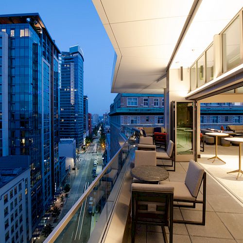 The image shows a modern rooftop terrace with seating, overlooking a cityscape with tall buildings during twilight.
