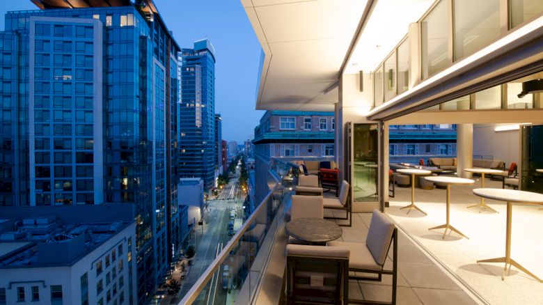 A modern rooftop patio with seating overlooks a city street lined with tall buildings, illuminated by evening lights.