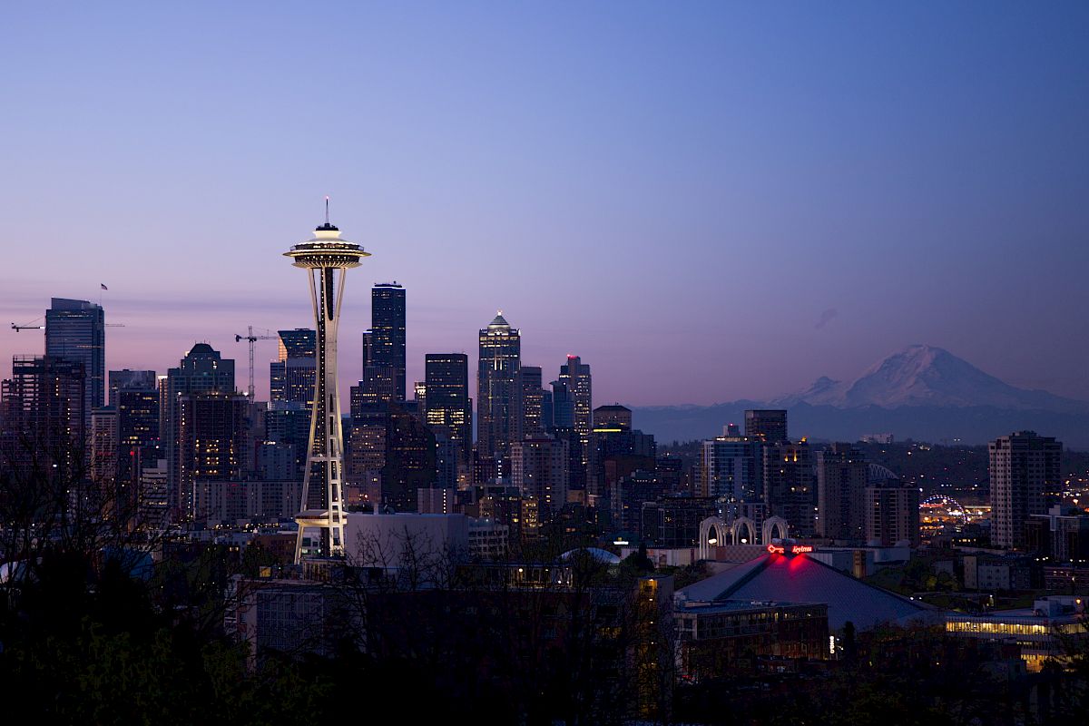 The image shows the Seattle skyline at dusk, featuring the Space Needle and Mount Rainier in the background under a purple sky.