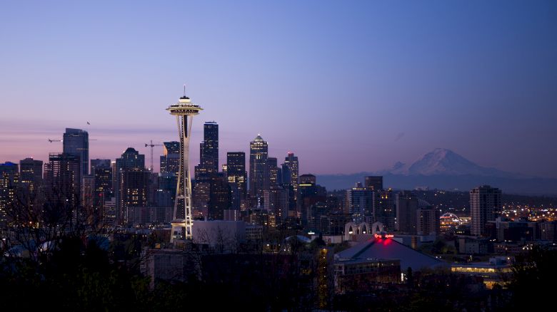 The image shows the Seattle skyline at dusk, featuring the Space Needle and Mount Rainier in the background under a purple sky.