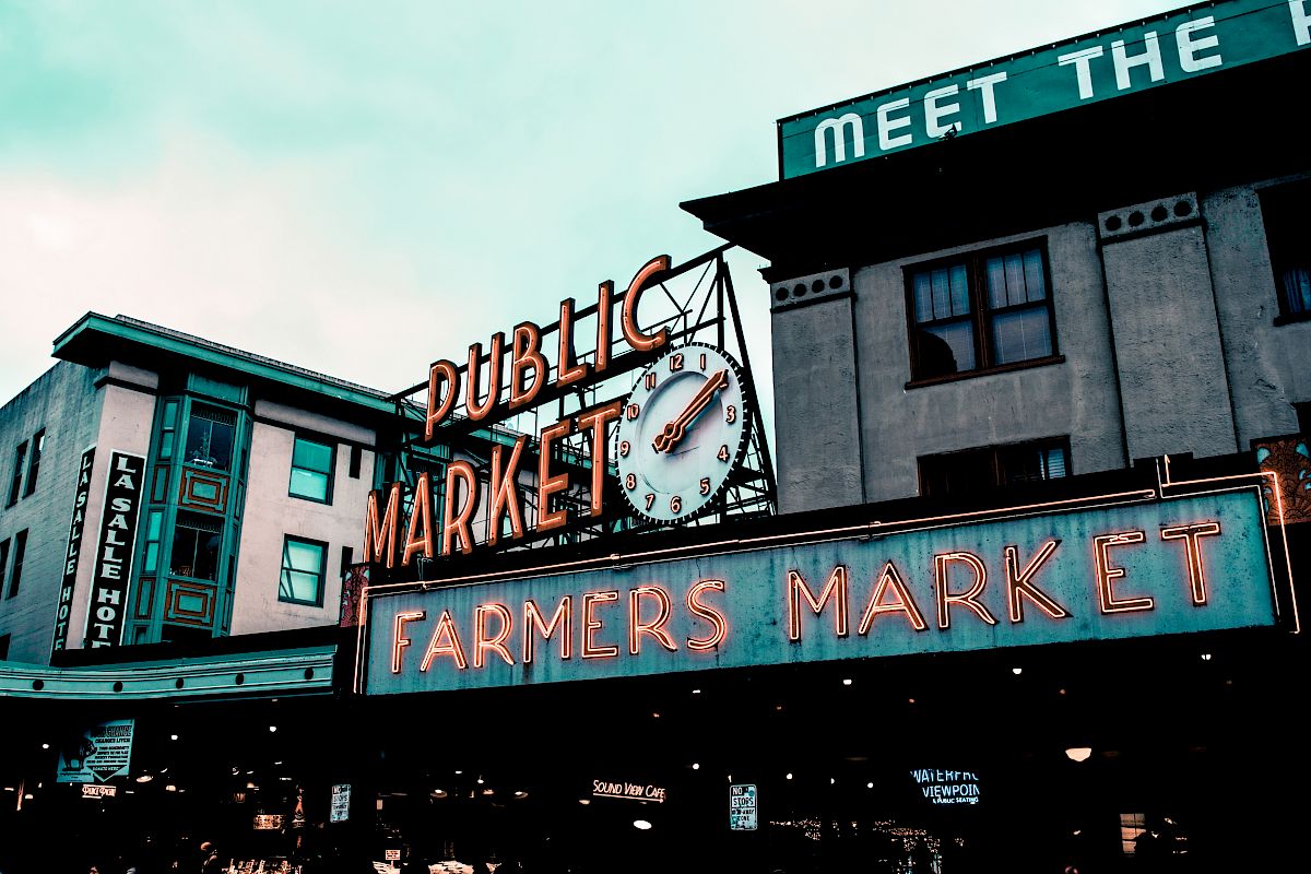Image shows a neon sign for a public market and farmers market, with buildings and a clock in the background.