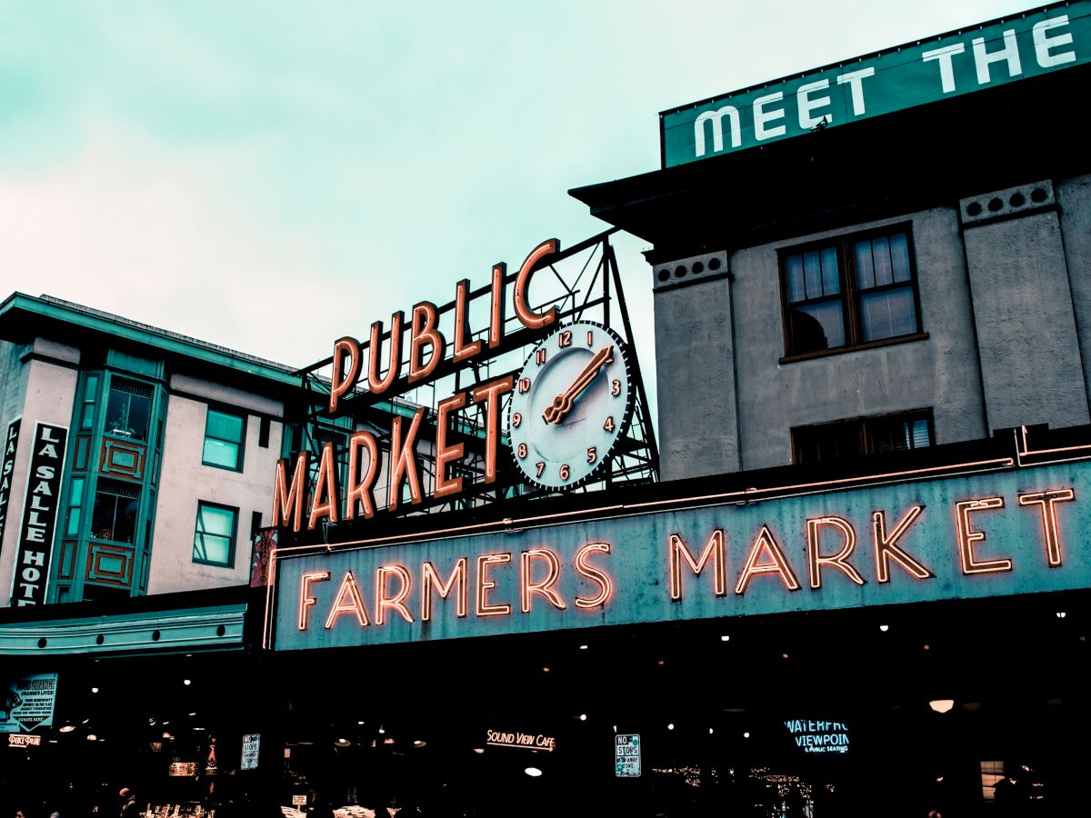 Image shows a neon sign for a public market and farmers market, with buildings and a clock in the background.