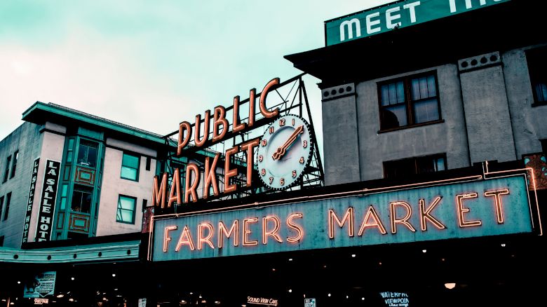 Image shows a neon sign for a public market and farmers market, with buildings and a clock in the background.