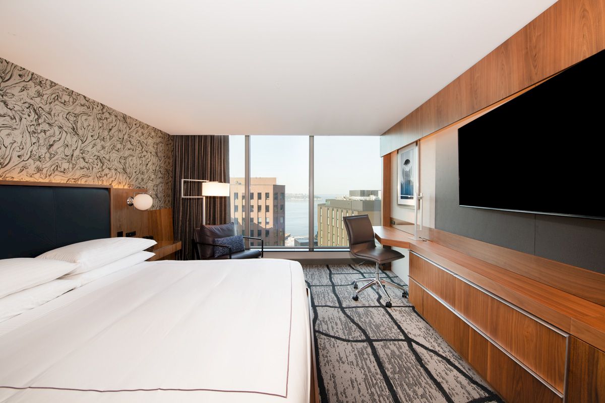 A modern hotel room with a large bed, a desk, a wall-mounted TV, and a window offering a city view.