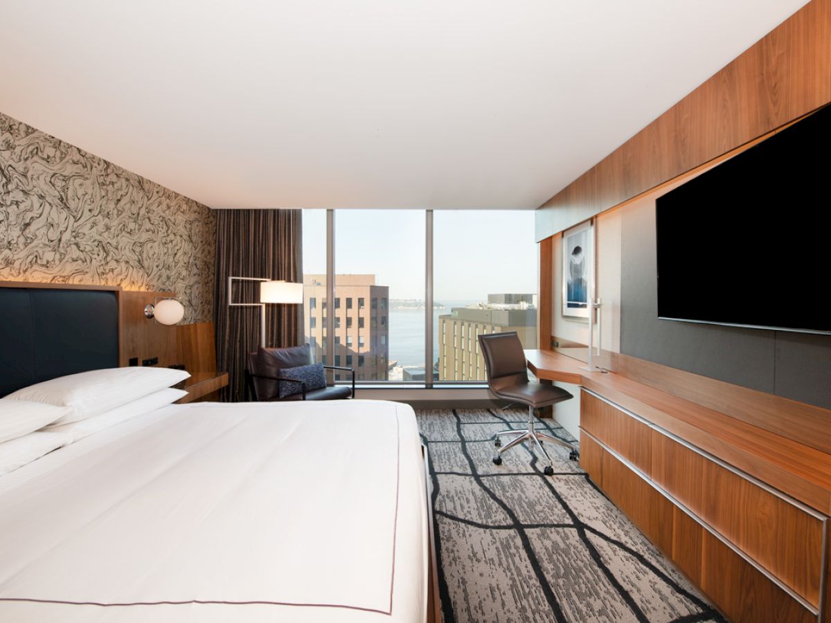 A modern hotel room with a large bed, a desk, a wall-mounted TV, and a window offering a city view.