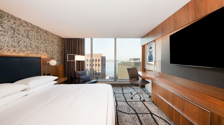 A modern hotel room with a large bed, a desk, a wall-mounted TV, and a window offering a city view.
