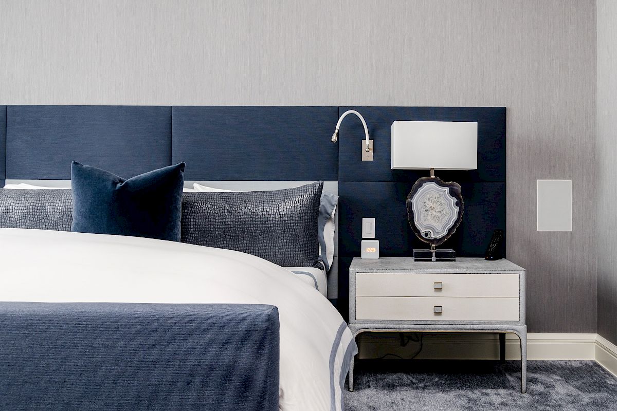 A modern bedroom with a navy-blue headboard, white nightstand, lamp, and textured bedding on a grey carpet.