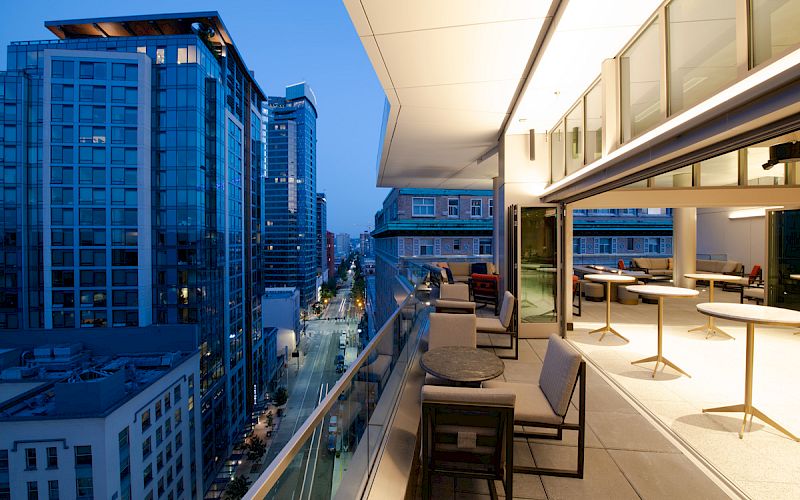 The image shows a modern cityscape at dusk with tall buildings and an outdoor terrace featuring seating and tables.