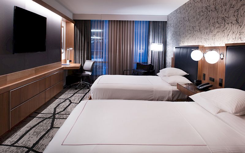 The image shows a modern hotel room with two beds, a wall-mounted TV, a desk, and stylish lighting and decor.