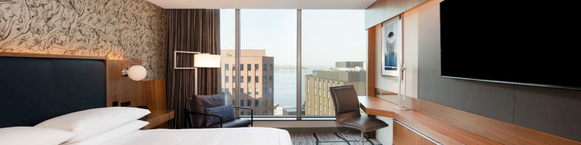 A modern hotel room with a king-sized bed, large TV, and a desk by the window overlooking city buildings.